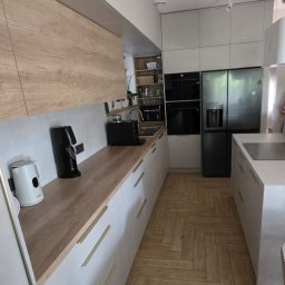 MEBLEGO - Nowoczesna kuchnia z drewnianym blatem i białymi szafkami, parkiet ułożony w jodełkę. Widoczny ekspres do kawy i lodówka side-by-side.