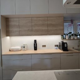 MEBLEGO - Jasna kuchnia z drewnianymi szafkami górnymi i białymi dolnymi. Na blacie ekspres do kawy, czajnik i saturator. W oknie storczyki. Minimalistyczny design.