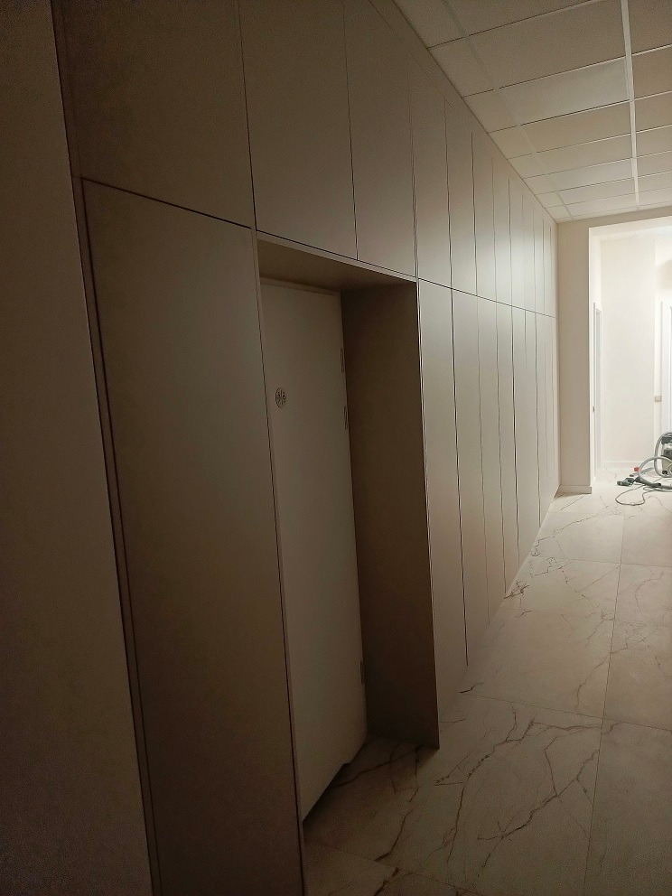 Długa ściana z szafami na wymiar w jasnym odcieniu, z ukrytymi drzwiami po lewej stronie, widoczne marmurowe płytki podłogowe i sufit kasetonowy, zdjęcie zrobione z niskiego kąta.