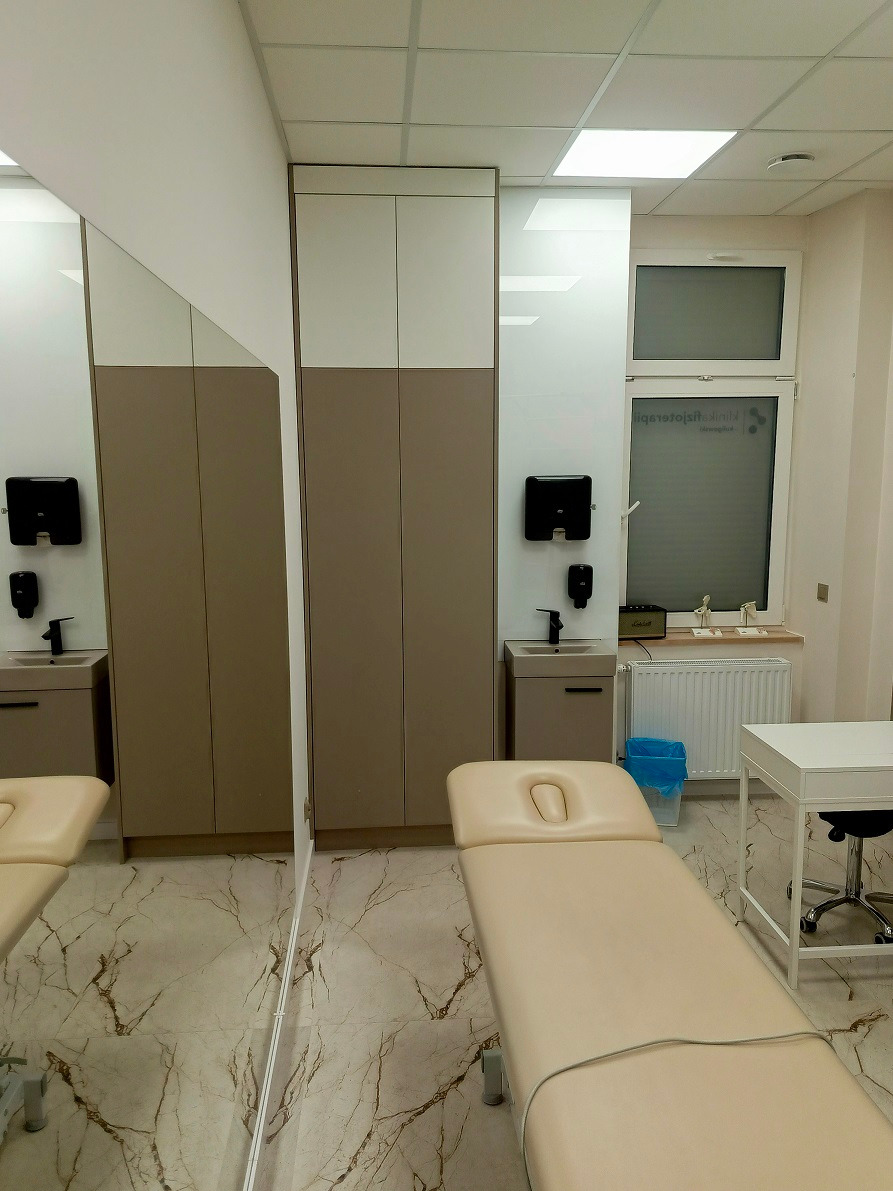 Beżowo-białe szafy na wymiar w gabinecie fizjoterapii, z umywalką i lustrem, widoczny stół do masażu, marmurowa podłoga.