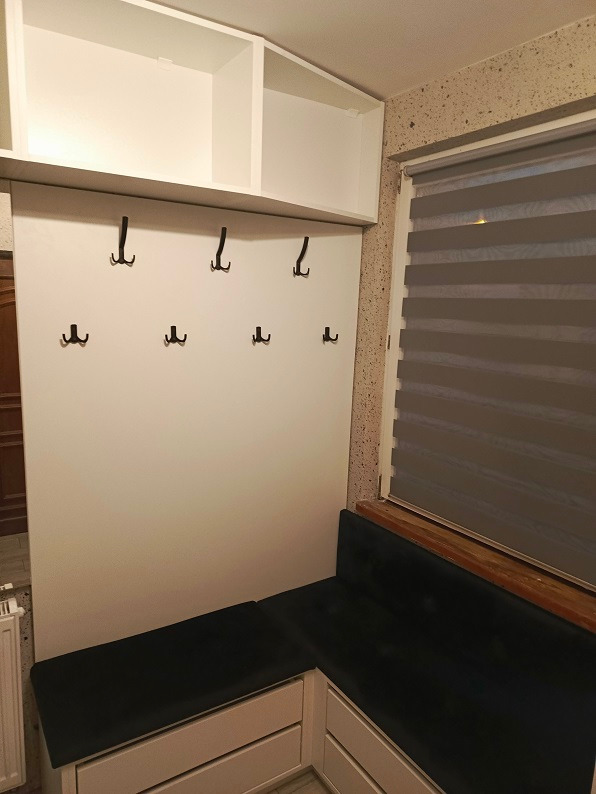 Biała zabudowa przedpokoju z szafkami wiszącymi, panelem z haczykami i siedziskiem z szufladami, uzupełniona o ciemnozieloną tapicerkę.