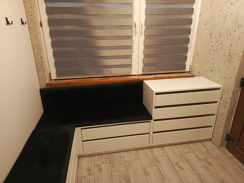 Biała zabudowa okienna z szufladami i tapicerowanym siedziskiem w kształcie litery L, z widocznymi haczykami na ścianie i żaluzjami w oknie.