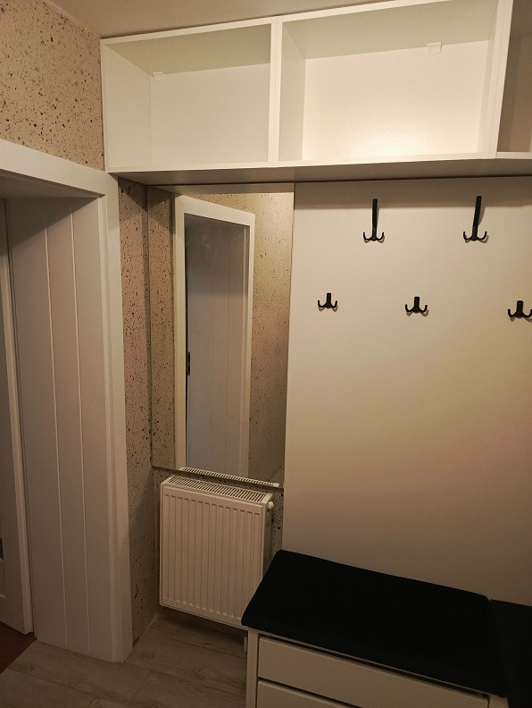 Przedpokój z białą zabudową na wymiar: szafki górne, panel z wieszakami, lustro nad grzejnikiem i siedzisko z szufladami.