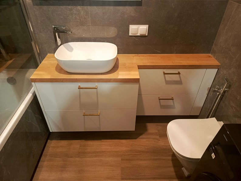 Nowoczesna łazienka z białą szafką pod umywalką z drewnianym blatem, umywalką nablatową, złotymi uchwytami, szarą płytką na ścianie i drewnopodobną podłogą.