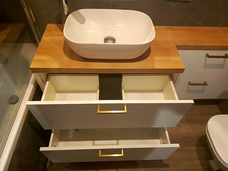 Łazienka z drewnianym blatem, umywalką nablatową i białymi szafkami z otwartymi szufladami z złotymi uchwytami.