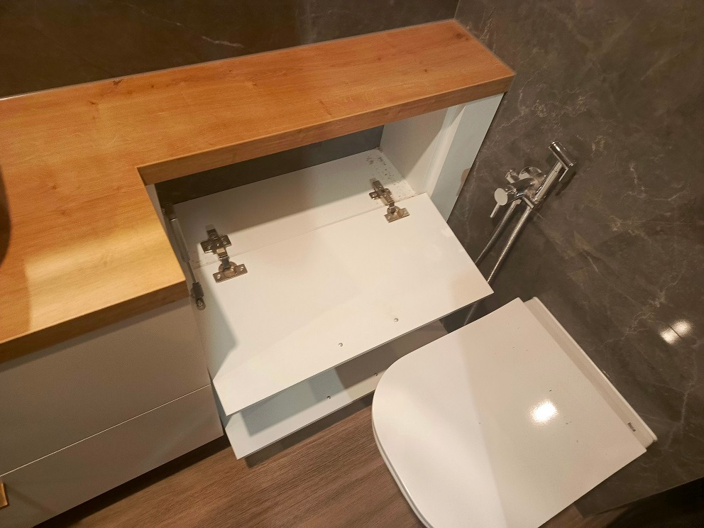 Biała szafka pod umywalkę z drewnianym blatem, otwarte drzwiczki ukazują zawiasy, obok widoczny bidet w łazience z marmurową ścianą.