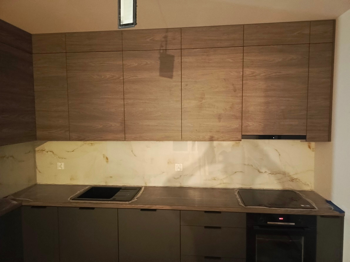 Nowoczesna kuchnia z górnymi szafkami w odcieniach drewna i szarymi dolnymi szafkami, marmurowy panel nad blatem, płyta indukcyjna i piekarnik elektryczny.