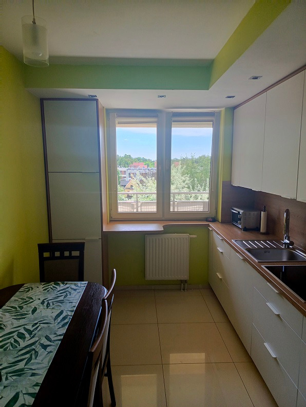 Jasna kuchnia z białymi szafkami i drewnianym blatem, widok na zewnątrz przez okno, stół z wzorzystym obrusem w lewym dolnym rogu.