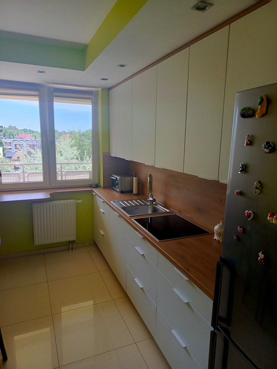 Jasna kuchnia z białymi frontami szafek, drewnianym blatem i widokiem na zieleń za oknem, lodówka ozdobiona magnesami.