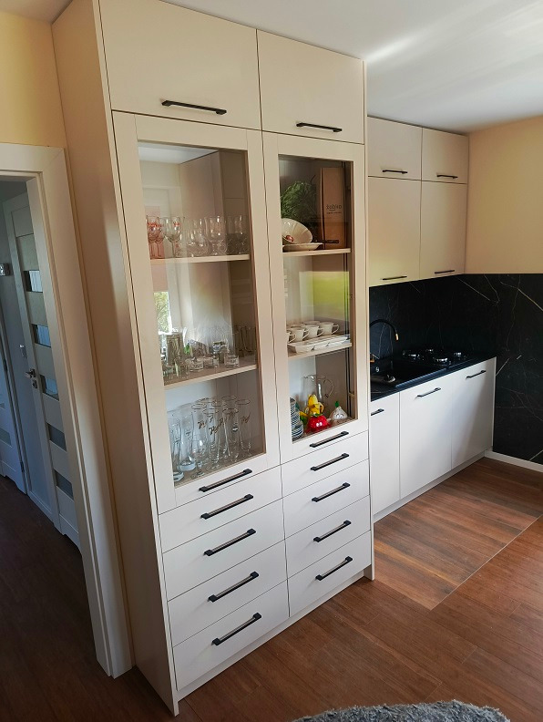 Jasne meble kuchenne z przeszkloną witryną i rzędem szuflad, czarne uchwyty, czarny blat z marmurkowym wzorem, drewniana podłoga.