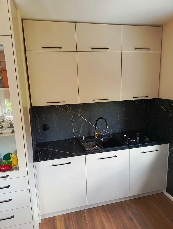 Jasna kuchnia z białymi frontami szafek i czarnym blatem imitującym marmur, złota bateria przy czarnym zlewozmywaku, widoczna płyta gazowa.