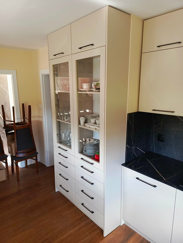 Biała, zabudowana szafa kuchenna z przeszkloną witryną, czarnymi uchwytami i blatem z czarnego marmuru, widok z boku na drewnianą podłogę i krzesła w tle.