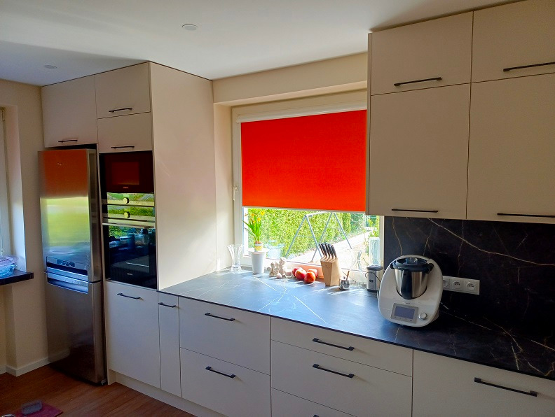 Jasna kuchnia z zabudową meblową, czarnym blatem imitującym marmur, czerwona roleta w oknie i sprzęt AGD, w tym Thermomix.