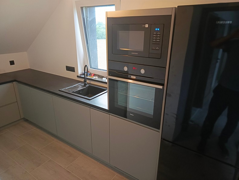 Nowoczesna kuchnia w szarościach z ciemnym blatem, zintegrowana zabudowa z piekarnikiem i mikrofalówką Amica oraz czarną lodówką Samsung, widok na okno dachowe.