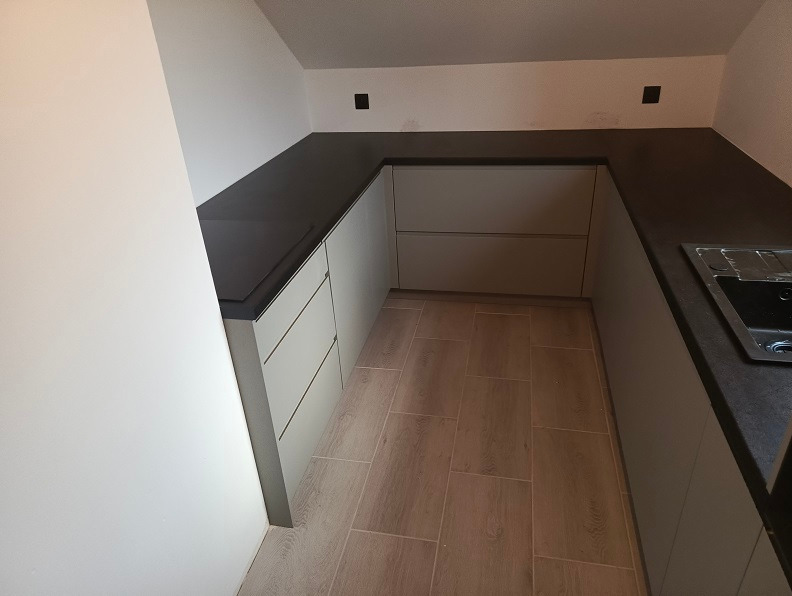 Nowoczesna kuchnia w odcieniach szarości z ciemnym blatem, minimalistyczne fronty szafek bez uchwytów, drewnopodobna podłoga, widok z perspektywy narożnika.