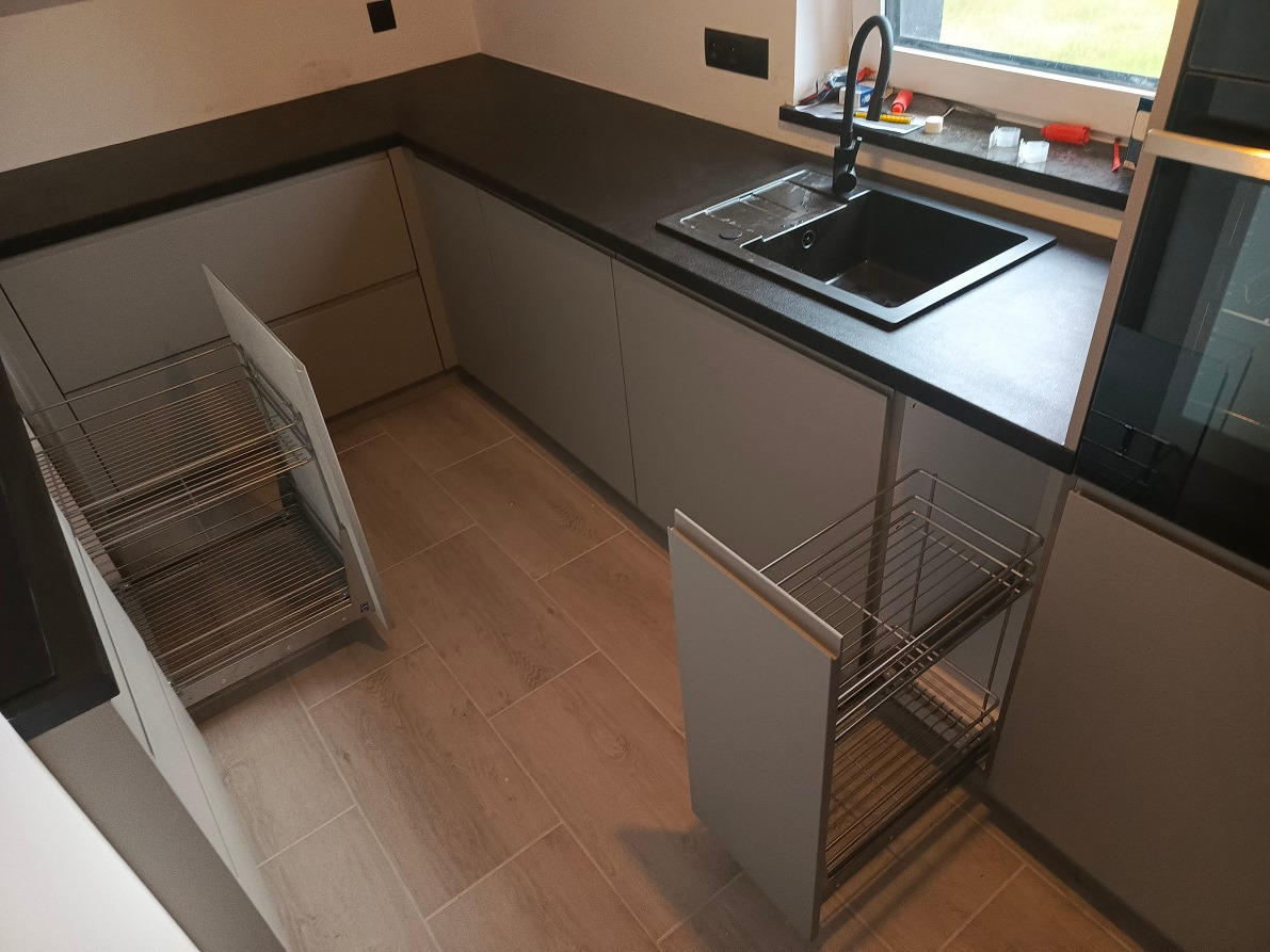 Nowoczesna kuchnia z szarymi frontami i czarnym blatem, wyposażona w wysuwane kosze cargo, widoczne płytki podłogowe imitujące drewno.