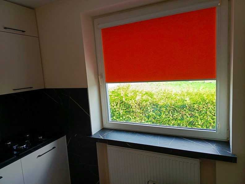 Fragment kuchni z białymi szafkami, czarnym blatem imitującym marmur i oknem z pomarańczową roletą, widok na zieleń za oknem.