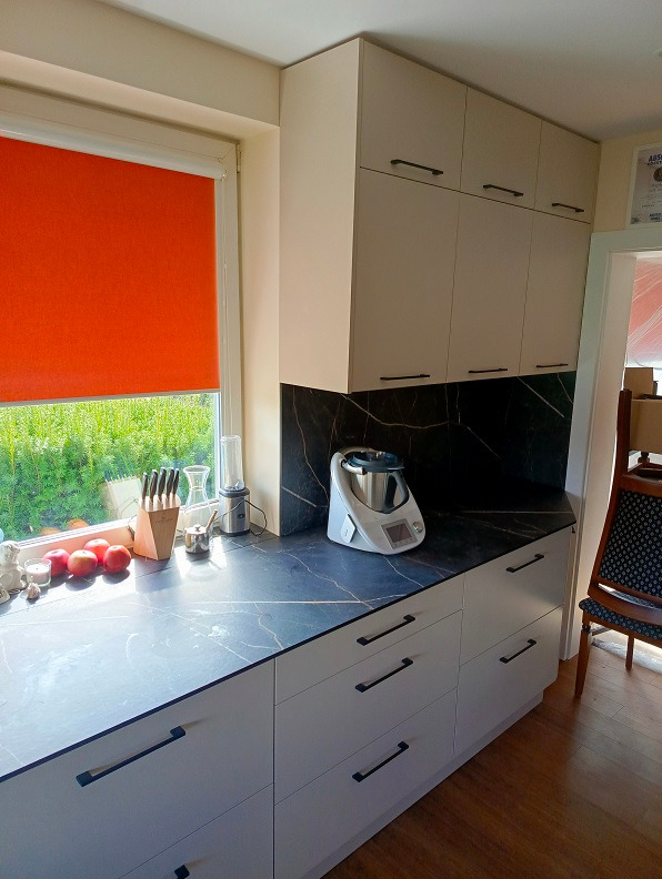 Jasna kuchnia z frontami bez uchwytów, ciemny blat z marmurowym wzorem, pomarańczowa roleta w oknie, widok na zieleń za oknem, na blacie naczynia i robot kuchenny.