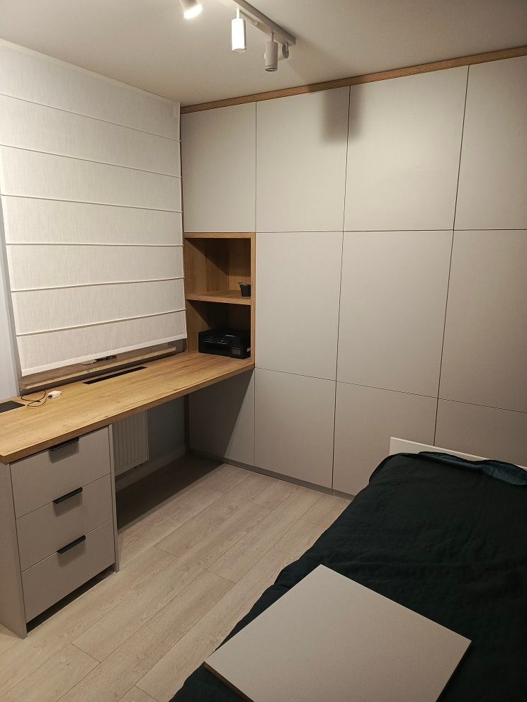 Szara zabudowa meblowa z biurkiem z drewnianym blatem, szufladami i otwartą półką, dopasowana do wnętrza sypialni z łóżkiem.