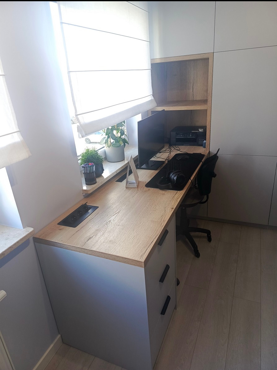 Biurko z blatem drewnianym wkomponowane w zabudowę meblową, z roślinami na parapecie i wbudowanym gniazdkiem elektrycznym.