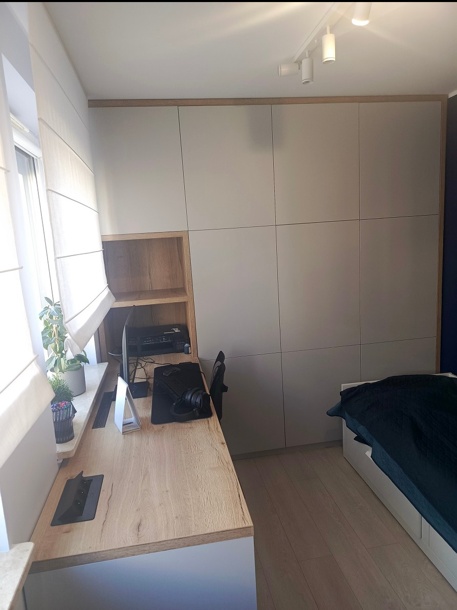 Szara szafa wnękowa z drewnianą ramą i otwartą półką, zintegrowana z biurkiem w pokoju z oknem i łóżkiem.