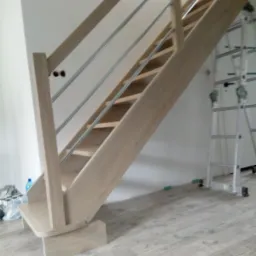 Jasne drewniane schody z metalowymi poręczami w trakcie montażu, widoczna drabina i butelka wody w tle.