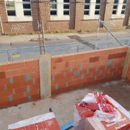 Wznoszone ściany domu z czerwonej cegły Porotherm, widoczne zbrojenia i zaprawa, w tle budynek z cegły z graffiti.