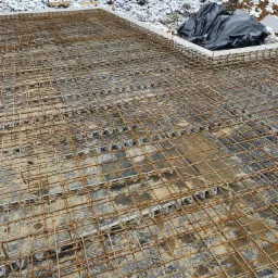 Zbrojenie fundamentu z siatki stalowej, częściowo przykryte czarną folią, na tle ośnieżonego terenu budowy i domów w oddali.