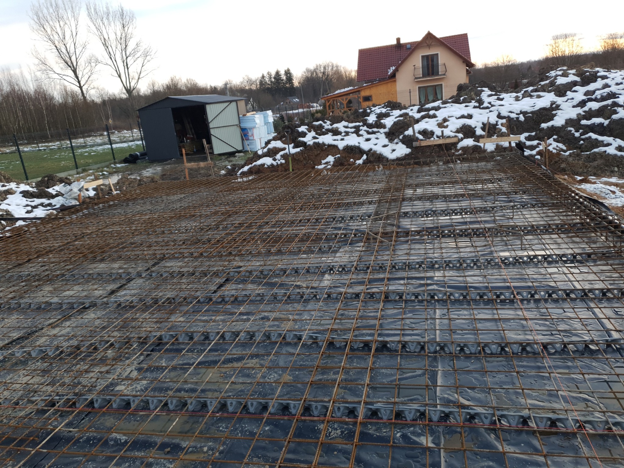 Zbrojenie fundamentu na czarnej folii z dystansami, widok na działkę z budynkiem w tle, częściowo pokrytą śniegiem.