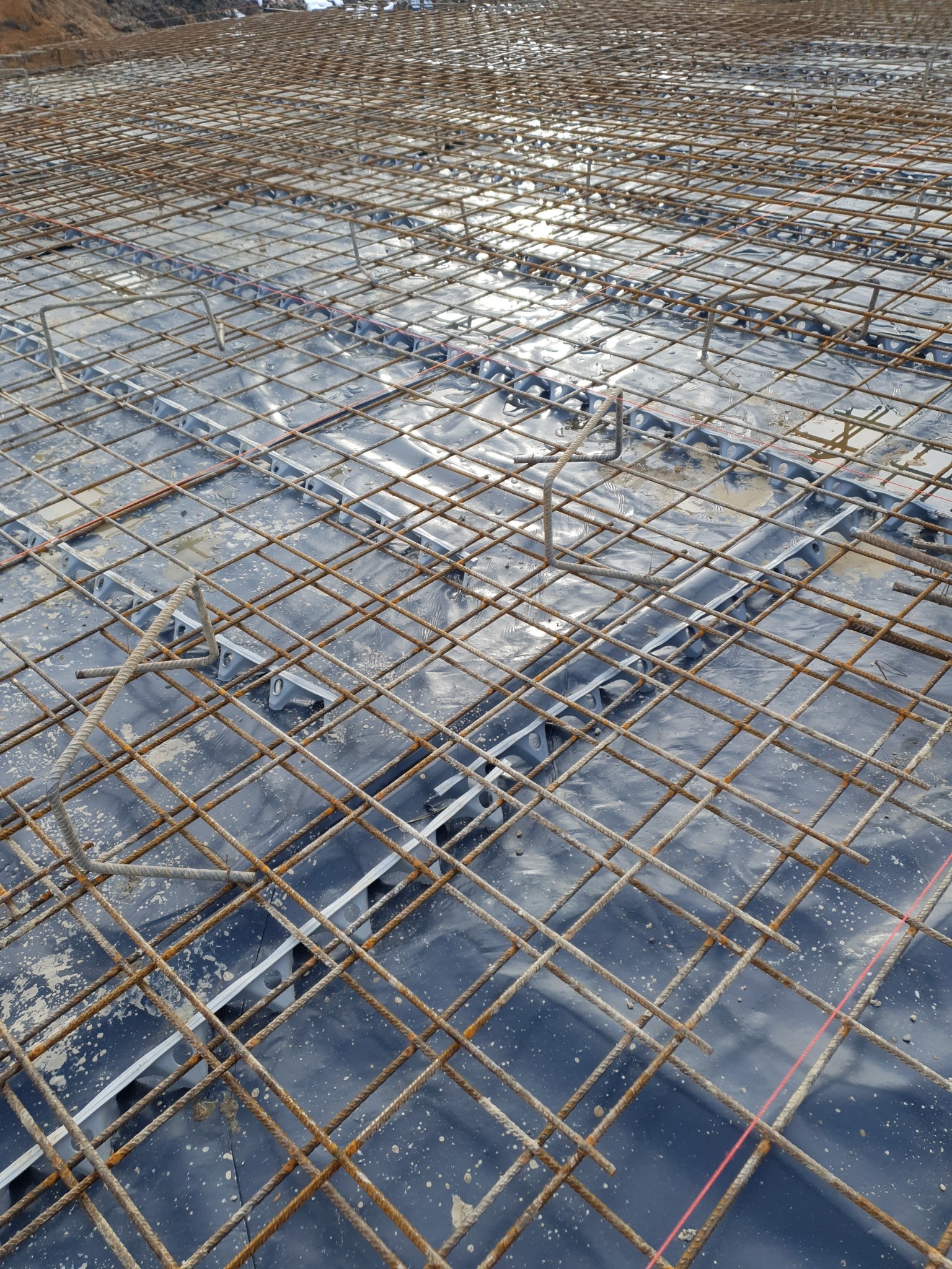 Zbrojenie płyty fundamentowej z siatki stalowej na folii hydroizolacyjnej, z widocznymi elementami dystansowymi i śladami betonu.