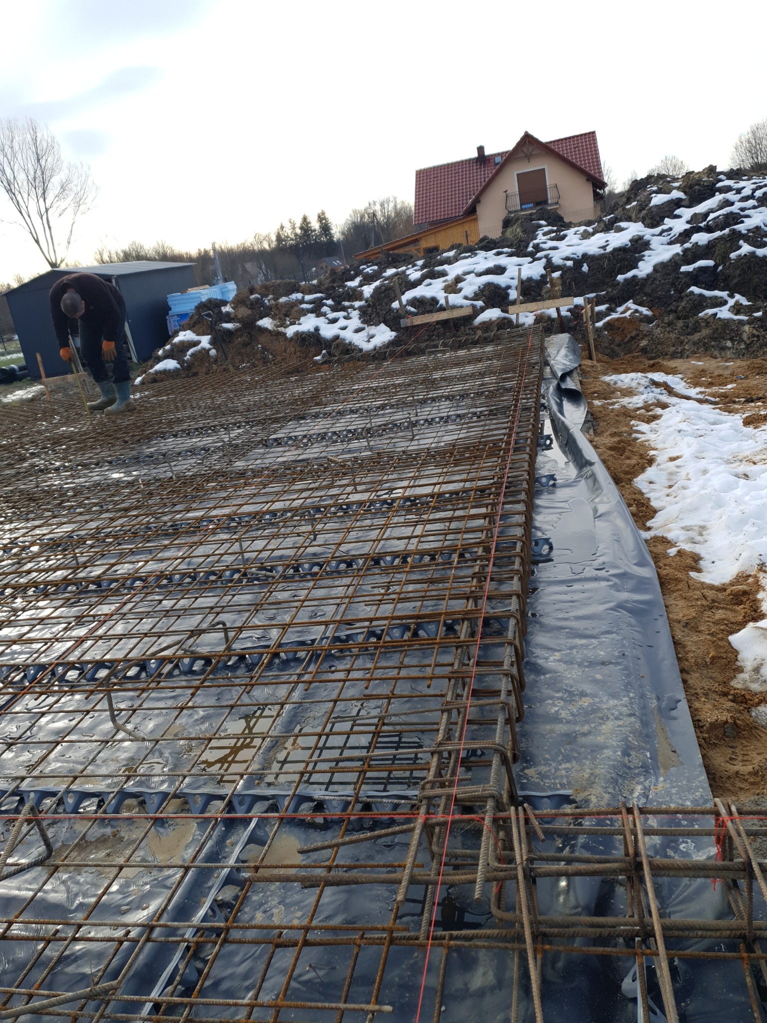 Zbrojenie fundamentu na folii, widoczne dystanse pod zbrojeniem, w tle pracownik w kaloszach i częściowo ośnieżony teren z budynkiem.