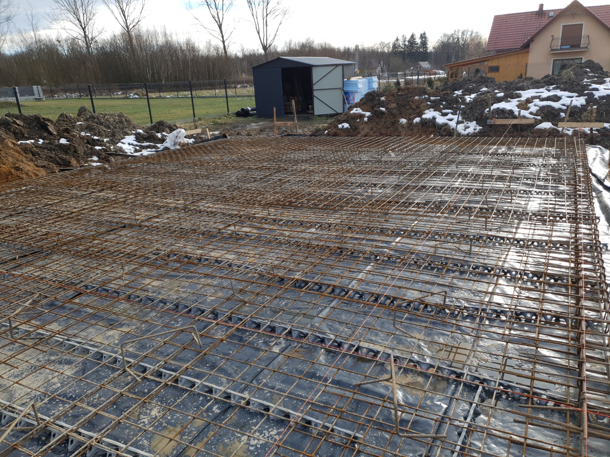 Siatka zbrojeniowa na folii, przygotowana do wylania fundamentów, widoczny teren budowy z ziemią i stertą śniegu w tle.