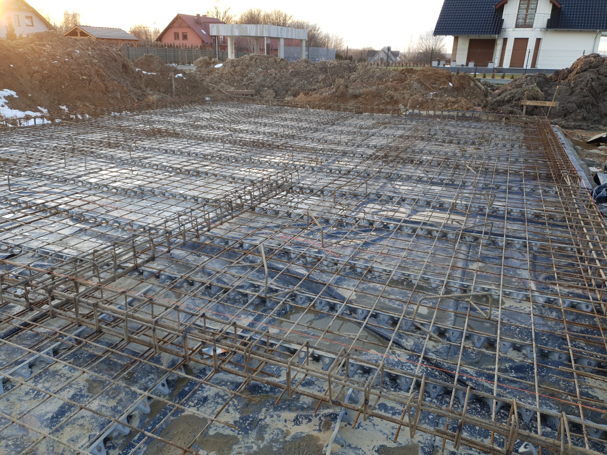Zbrojenie fundamentu z siatki stalowej na podkładzie z folii i dystansów, widoczne przygotowane wykopy i dom w tle.