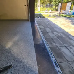 Szara posadzka w garażu z odpływem liniowym przy wyjeździe, widok na zewnątrz z drzewami i koszem do koszykówki.