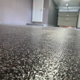 Szara posadzka żywiczna z drobnymi kolorowymi wtrąceniami w garażu, widoczna brama garażowa i białe drzwi wejściowe.