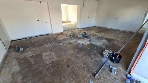 Przygotowanie podłoża w pustym pomieszczeniu przed wylaniem nowej posadzki, widoczne narzędzia, mieszadło, wiadro z farbą, szlifierka i kable elektryczne rozłożone na podłodze.