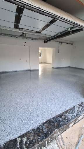 Gładka, jasnoszara posadzka z drobnymi, ciemnymi inkluzjami w garażu z otwartą bramą, widoczne elementy konstrukcyjne sufitu i wylewka przy krawędzi posadzki.