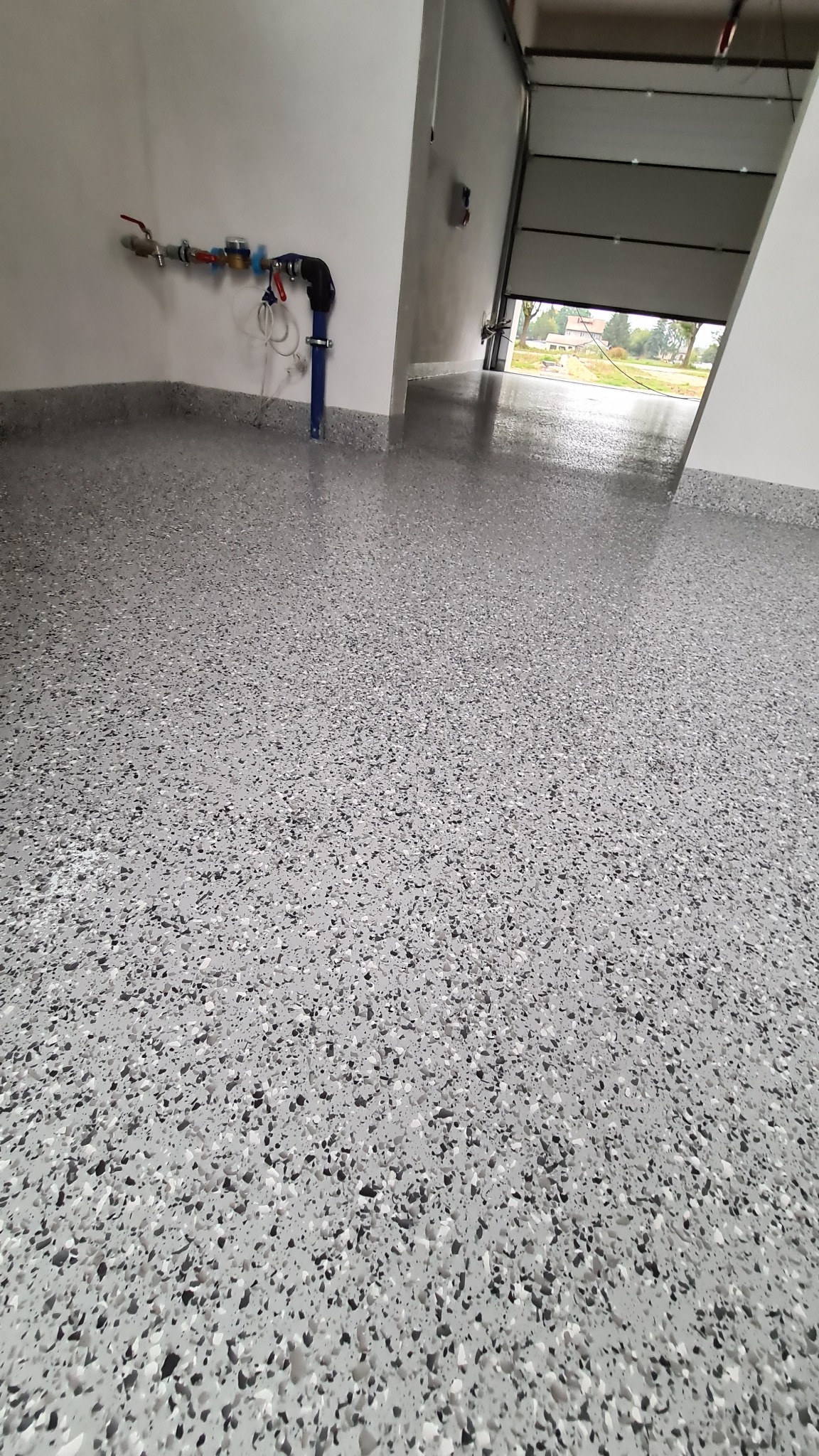 Jasnoszara posadzka z drobnymi, nieregularnymi plamkami w odcieniach szarości i czerni, widoczna instalacja wodna przy ścianie i brama garażowa z widokiem na zewnątrz.