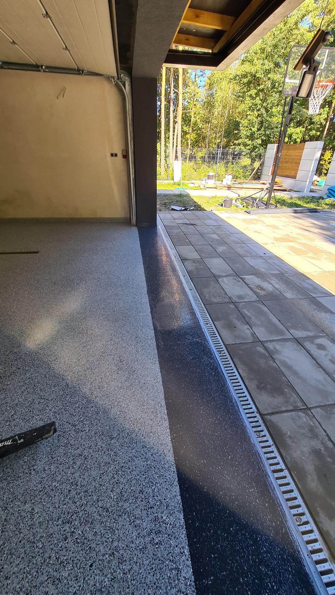 Szara posadzka w garażu z odpływem liniowym przy wyjeździe, widok na zewnątrz z drzewami i koszem do koszykówki.