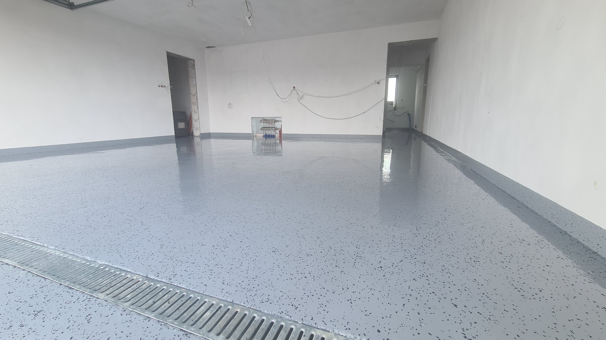 Świeżo wylana, szara posadzka żywiczna z ciemnymi drobinkami w surowym wnętrzu, widoczna metalowa kratka odpływowa przy ścianie, instalacja grzewcza na ścianie w tle.