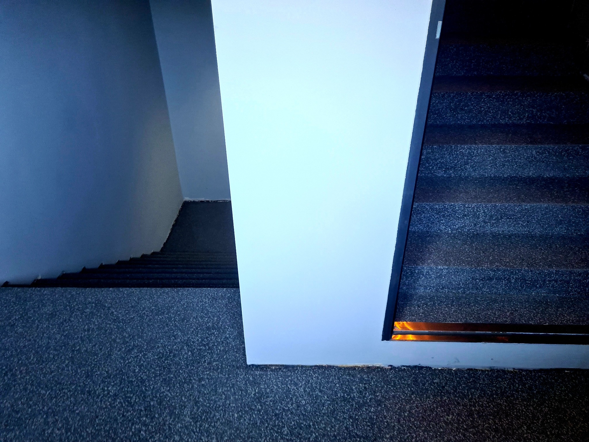 Szare schody z antypoślizgową, teksturowaną nawierzchnią i metalowymi listwami zabezpieczającymi krawędzie stopni, widoczne z góry, z fragmentem ściany pomalowanej na szaro i białym filarem po...