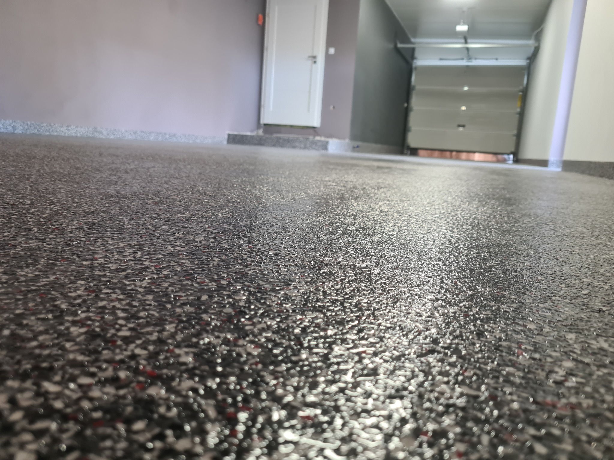 Szara posadzka żywiczna z drobnymi kolorowymi wtrąceniami w garażu, widoczna brama garażowa i białe drzwi wejściowe.