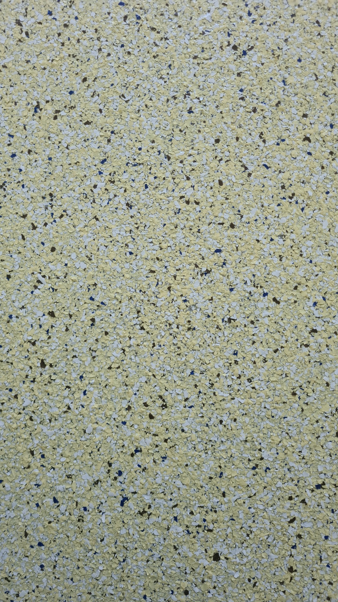 Jasnożółta posadzka żywiczna z ciemnymi i niebieskimi drobinami, widok z góry, równomierna tekstura.