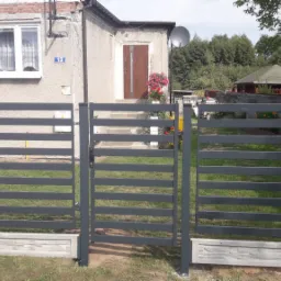 Szare, poziome przęsła ogrodzenia z furtką na betonowej podmurówce, na tle domu z kwiatami w doniczce przy wejściu.