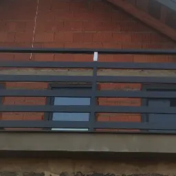 Szara, metalowa balustrada balkonowa z poziomymi prętami, zamontowana na balkonie budynku z czerwonej cegły.