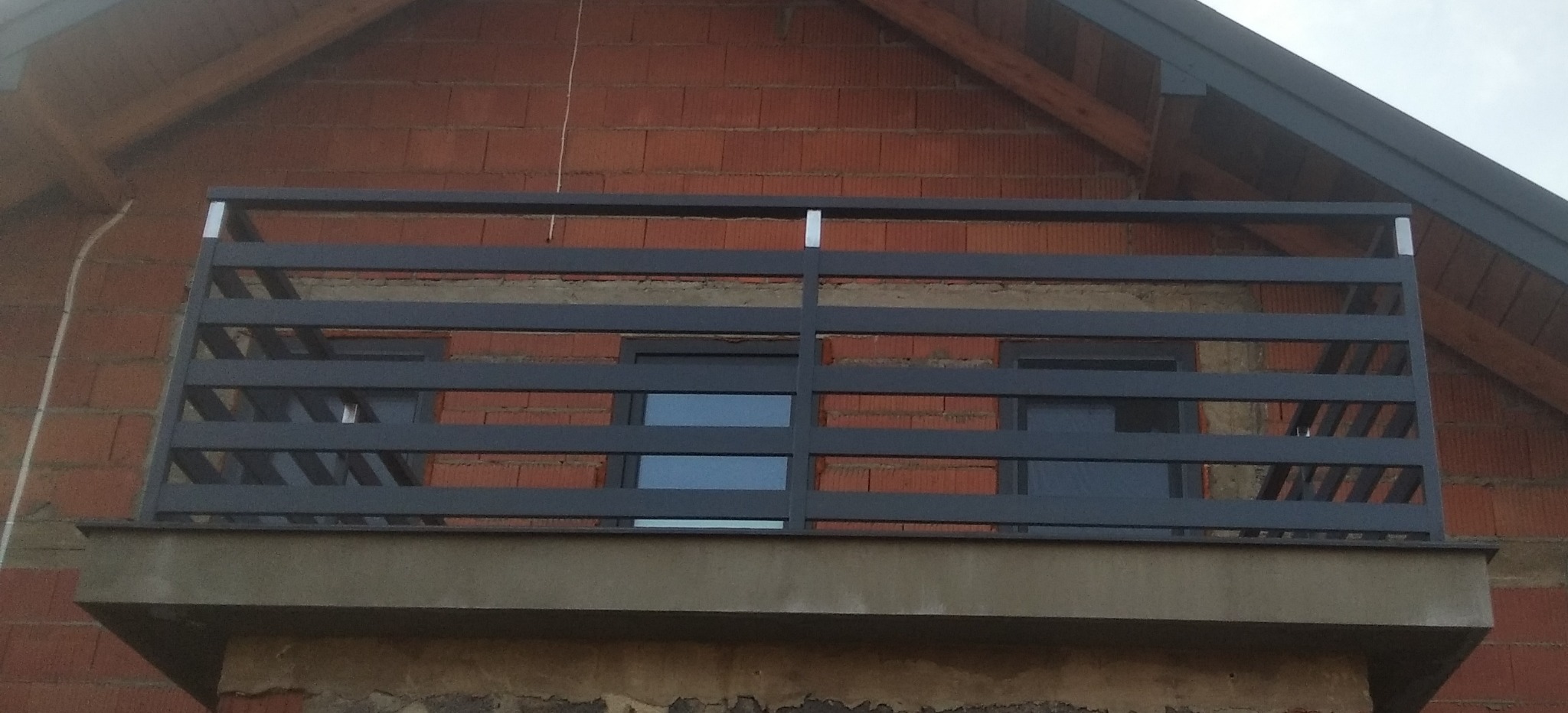 Szara, metalowa balustrada balkonowa z poziomymi prętami, zamontowana na balkonie budynku z czerwonej cegły.