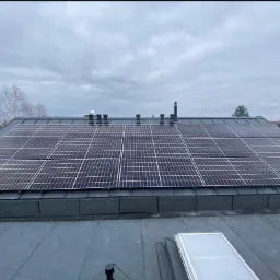 Panele słoneczne na dachu budynku z szarej blachy w pochmurny dzień, widoczne kominy i elementy wentylacyjne.