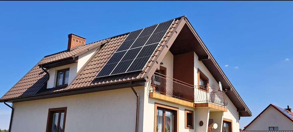 Dom jednorodzinny z brązowym dachem, na którym zamontowano panele słoneczne; widoczny komin i balkon z ozdobną balustradą.