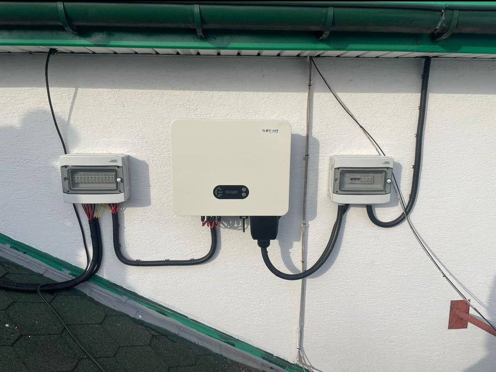 Biały falownik SOFAR zamontowany na elewacji budynku, po bokach dwie puszki elektryczne z okablowaniem w peszlach, widoczny fragment dachu pokrytego zieloną dachówką.