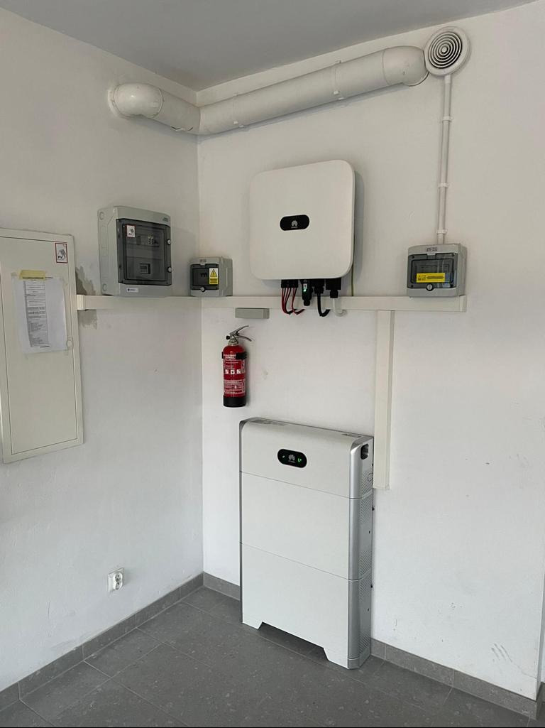 Wnętrze pomieszczenia technicznego z zamontowanym inwerterem i baterią energii, instalacją elektryczną, wentylacją oraz gaśnicą.